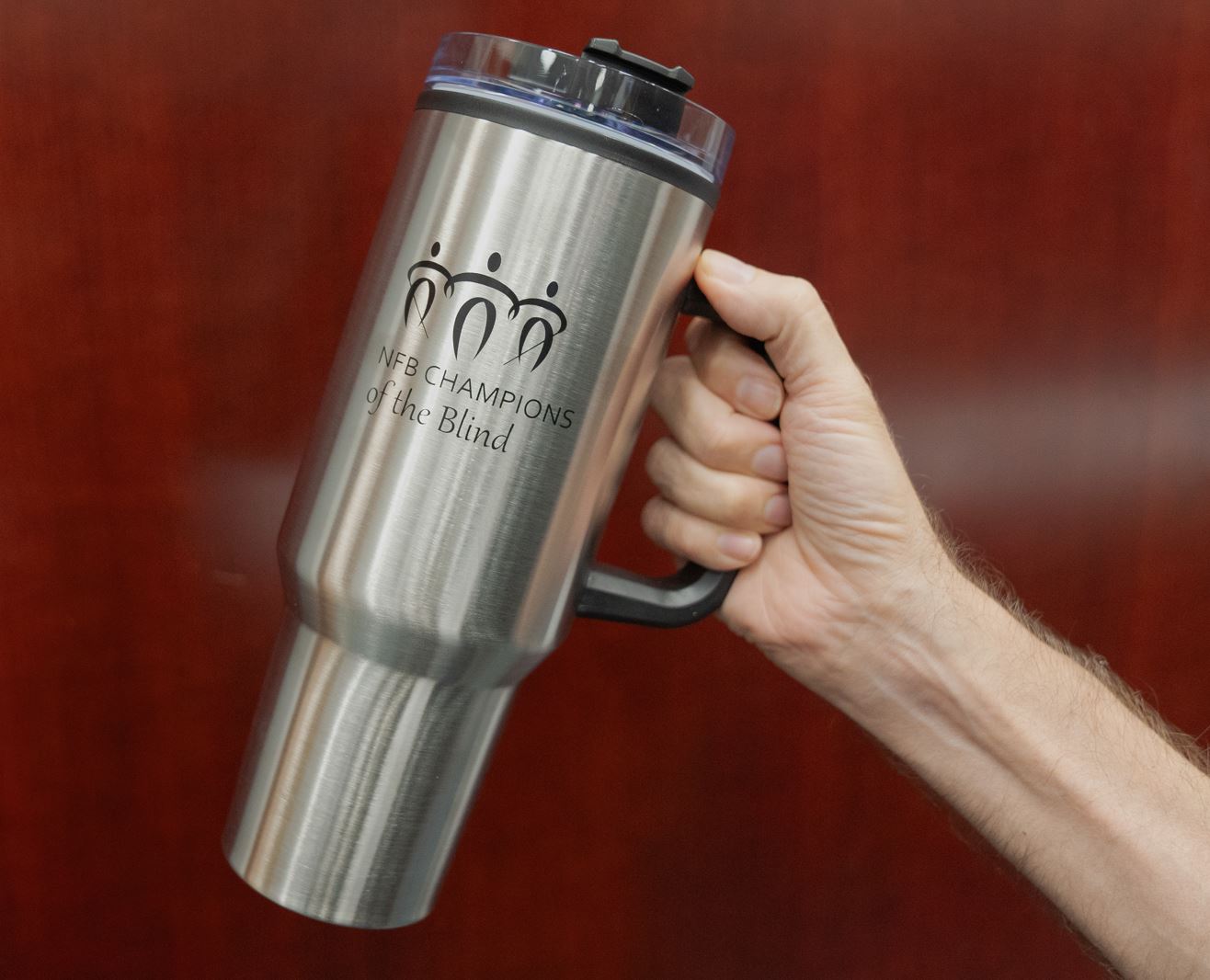 A persons hand holding a large insulated metallic mug against a rich wood background. On the mug is a stylized graphic of three people linking arms, two are holding canes. Below the graphic is printed NFB Champions of the Blind in black lettering