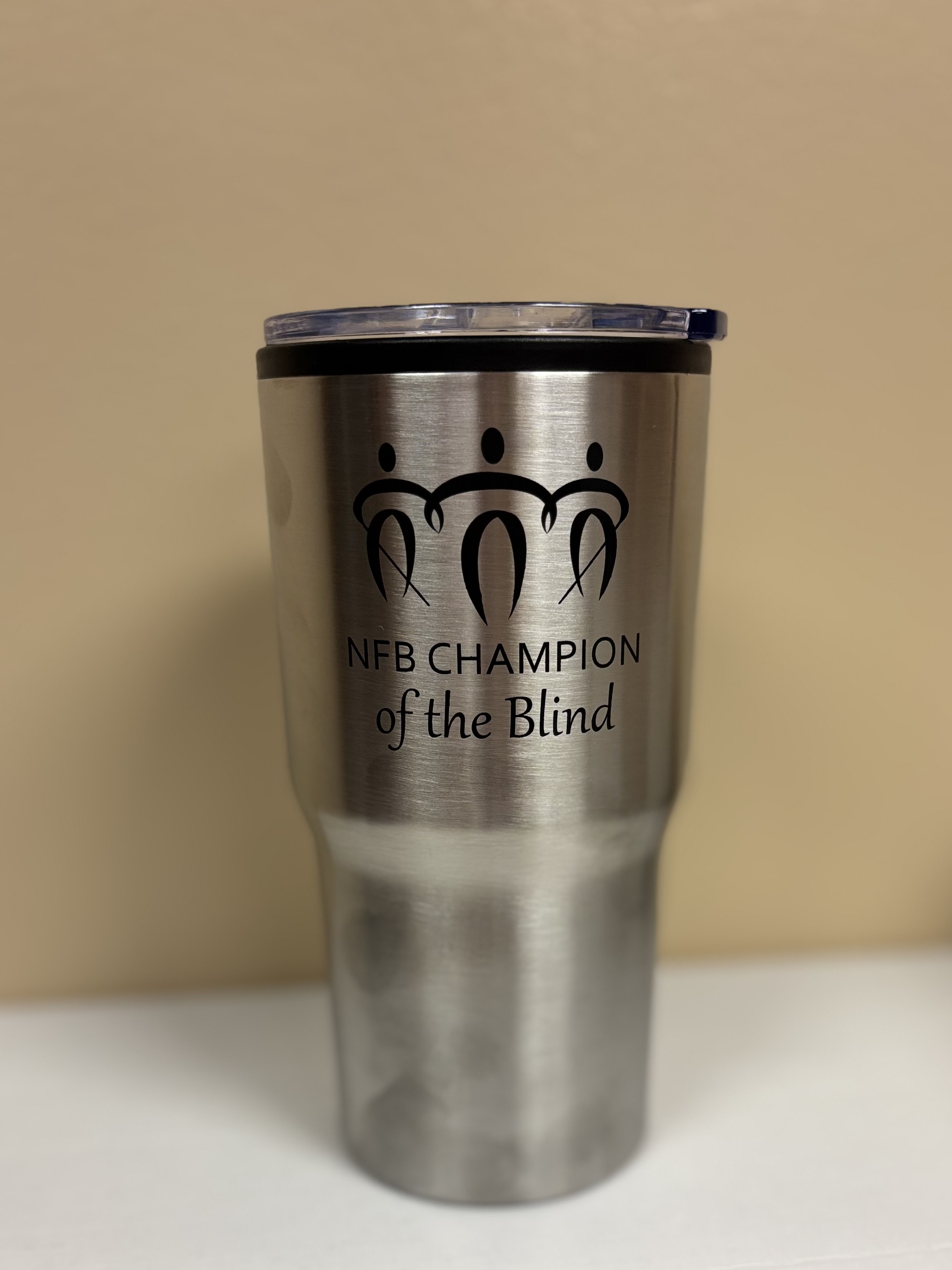 A large insulated metallic mug against a tan background. On the mug is a stylized graphic of three people linking arms, two are holding canes. Below the graphic is printed NFB Champion of the Blind in black lettering